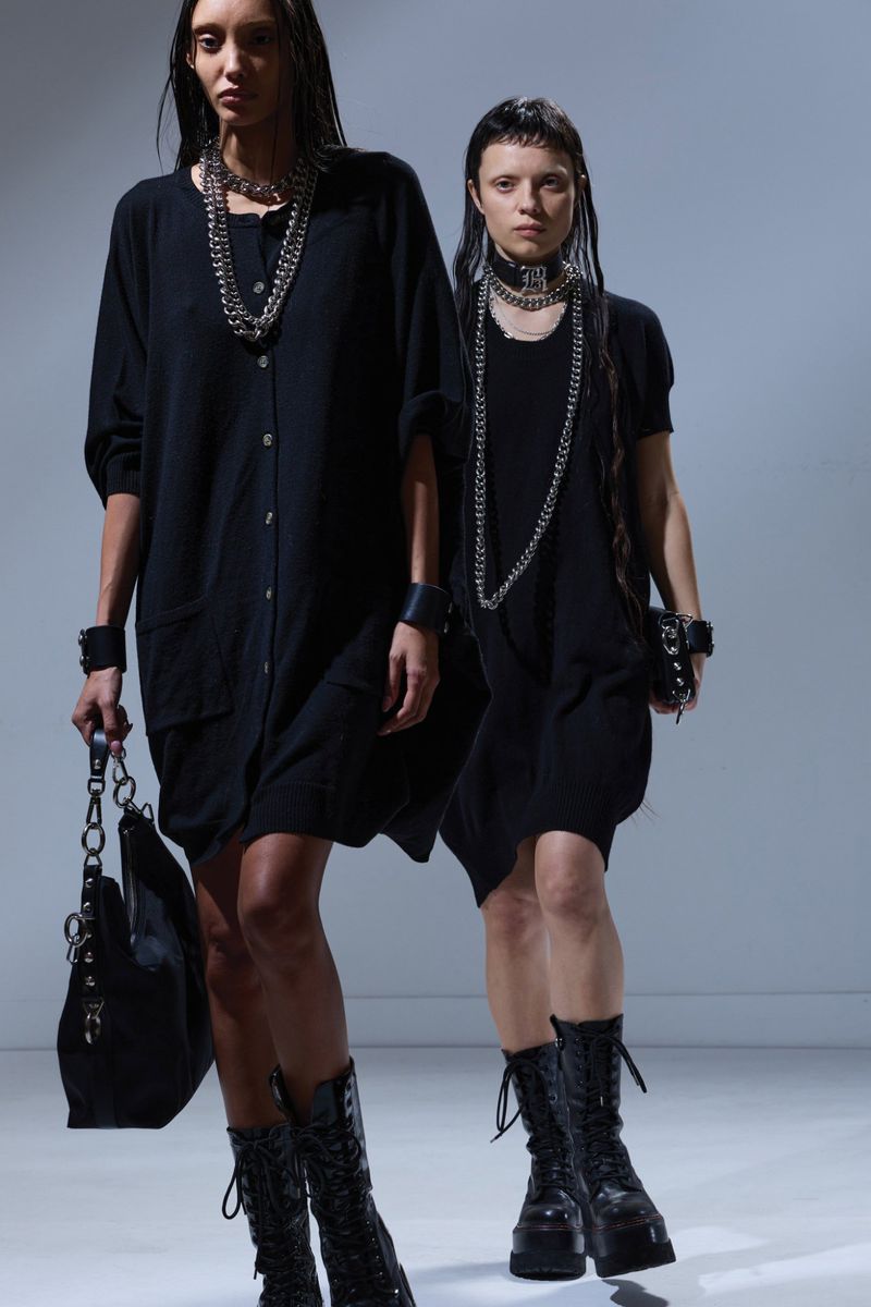 Two women in black dresses and heavy necklaces pose in a studio with dark boots and matching bracelets or cuffs, fashionable and edgy.