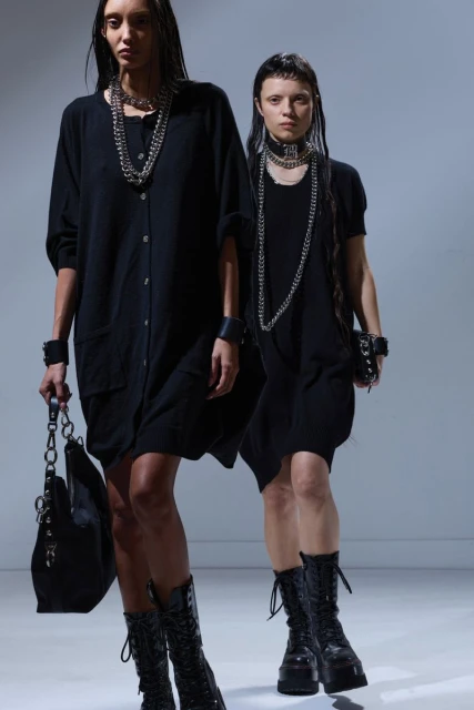 Two women in black dresses and heavy necklaces pose in a studio with dark boots and matching bracelets or cuffs, fashionable and edgy.