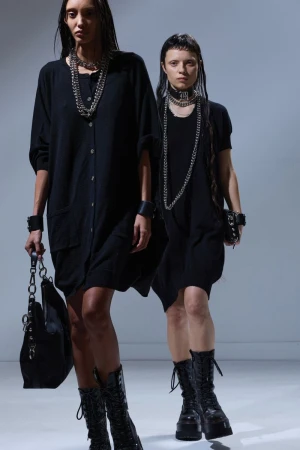 Two women in black dresses and heavy necklaces pose in a studio with dark boots and matching bracelets or cuffs, fashionable and edgy.