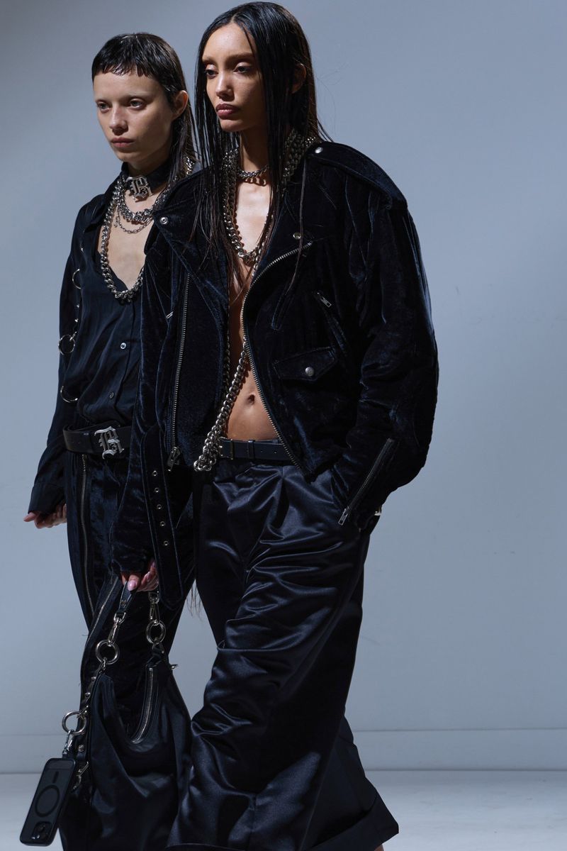 Two fashion models present avant-garde apparel. One dons a velvet motorcycle jacket and silk pants, while the other wears a dark jumpsuit.