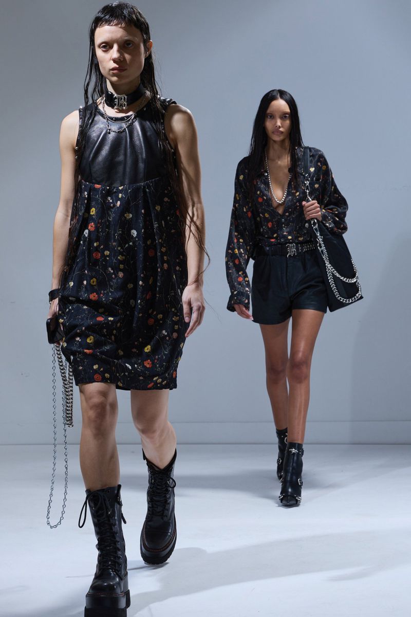 Two fashion models walk a runway in black leather-accented dresses and combat boots during a fashion show against a grey background.
