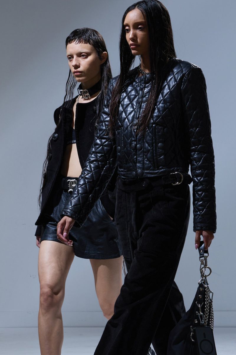 Two women in black outfits pose in a fashion studio, showcasing dark fashion, leather, and unique hairstyles against a neutral backdrop.
