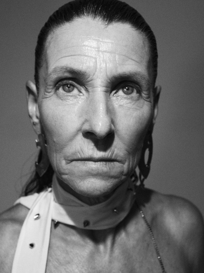 Close-up monochrome portrait of an elderly woman with slicked-back dark hair wearing a white neck scarf and dark earrings