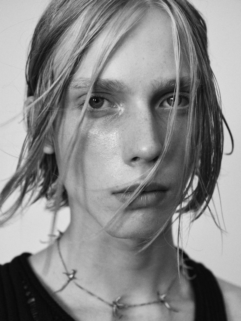 Close-up portrait of a young model with blonde hair falling across face wearing a dark top and necklace against a light background