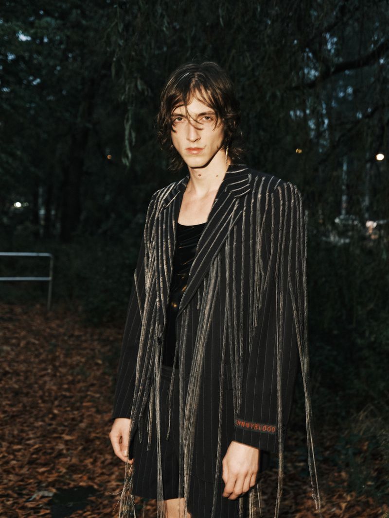 Adult male with long hair poses in an elegant black pinstripe jacket in a park, with a dark, shadowy background of trees and leaves.