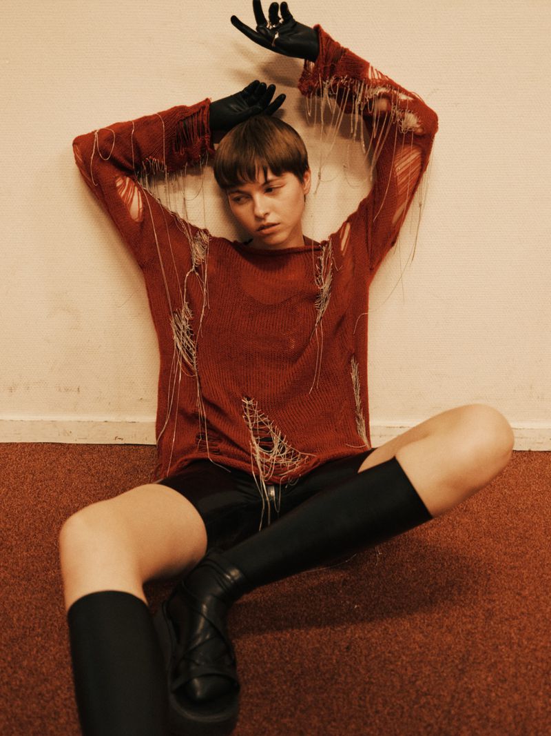 A person poses indoors wearing a distressed knit sweater with black gloves, shorts and boots, leaning against a cream-colored wall.