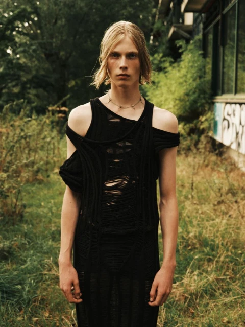 An adult male stands tall outdoors, showcasing a distressed black top and necklace, complemented by his fair hair and the overgrown surroundings.