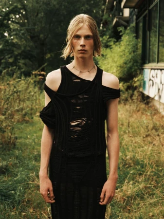 An adult male stands tall outdoors, showcasing a distressed black top and necklace, complemented by his fair hair and the overgrown surroundings.