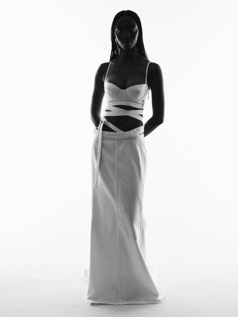 Black woman in white bra top and long skirt poses in a studio with high-key lighting, highlighting her silhouette and fashion.