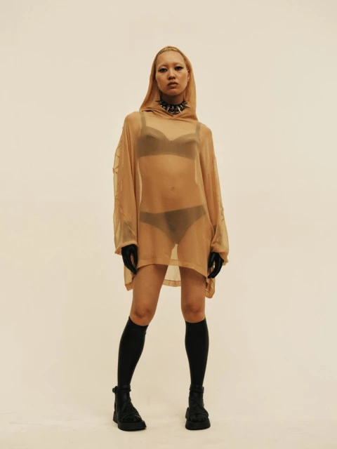 Adult woman poses in a semi-sheer hooded mini dress with black undergarments in a studio. She wears a choker, socks, and platform shoes.