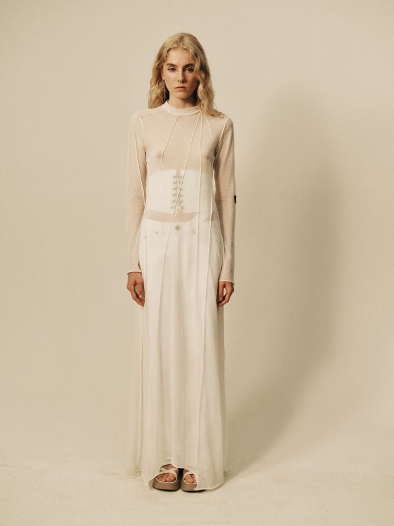 Full-length studio shot of a blonde woman in a sheer top, corset, and long white skirt, posing in front of a neutral background.