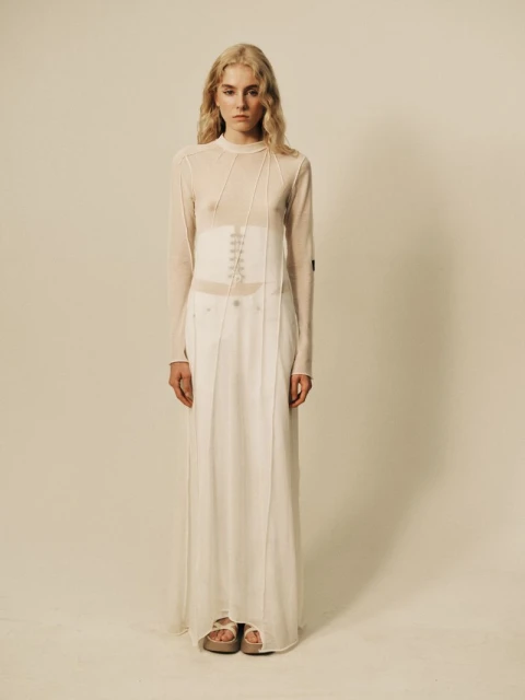 Full-length studio shot of a blonde woman in a sheer top, corset, and long white skirt, posing in front of a neutral background.