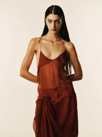 Model with long dark hair wearing a sheer rust red dress posing against a plain neutral background