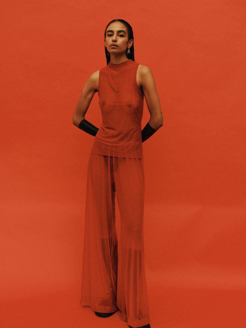 A woman poses in a sheer orange outfit against a matching backdrop, standing full-length with hands behind her back, showcasing fashion.