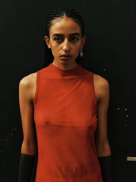 Portrait of an androgynous young adult with braided hair in a red sleeveless dress standing against a dark background in a studio setting.