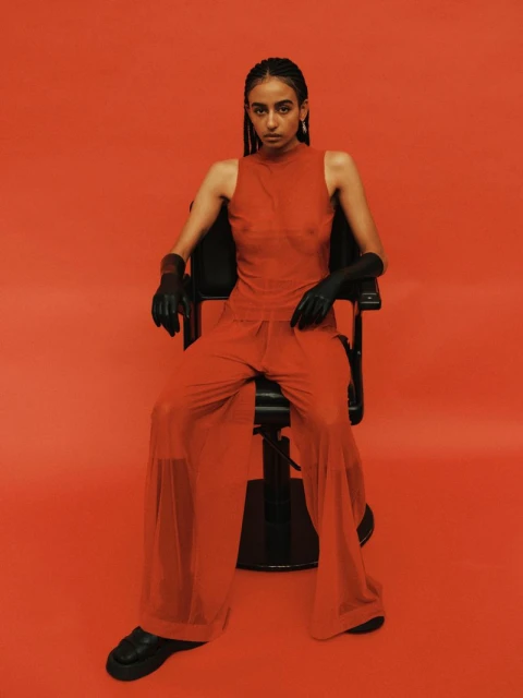 Portrait of an adult woman with braided hair, wearing a red outfit and black gloves, seated in a barber chair against a red background.
