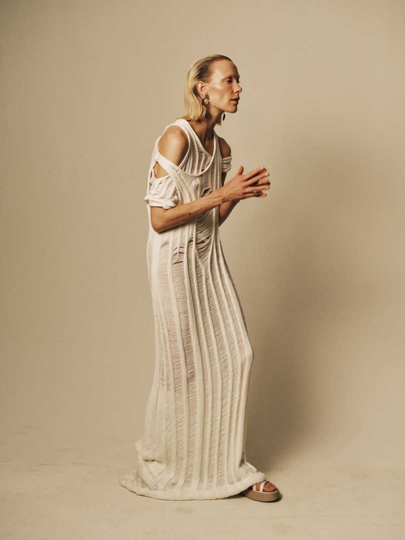 Adult woman in a long, cream-colored, shredded-knit dress and sandals stands indoors against a neutral backdrop, posing in profile.