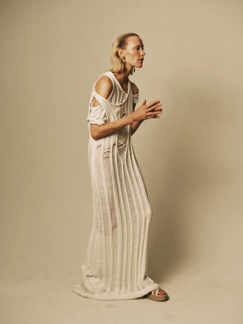 Adult woman in a long, cream-colored, shredded-knit dress and sandals stands indoors against a neutral backdrop, posing in profile.