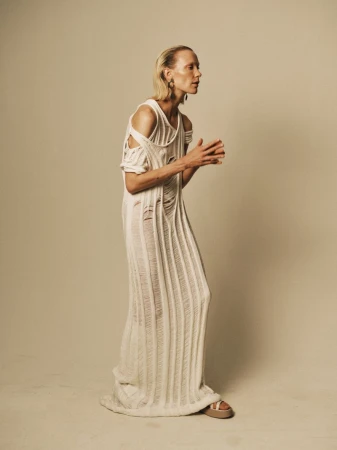 Adult woman in a long, cream-colored, shredded-knit dress and sandals stands indoors against a neutral backdrop, posing in profile.