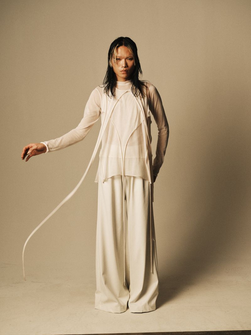 A slender person with wet hair stands in a photo studio, dressed in an all-white outfit with long, wide-leg pants and a sheer top.