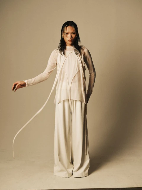A slender person with wet hair stands in a photo studio, dressed in an all-white outfit with long, wide-leg pants and a sheer top.