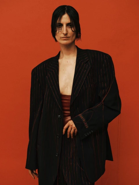 Woman in oversized pinstripe blazer and rust-colored crop top poses against an orange background in a fashion portrait. Studio lighting.