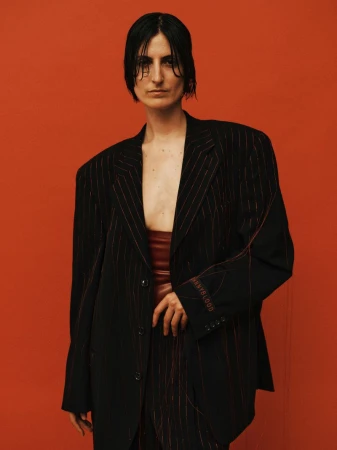 Woman in oversized pinstripe blazer and rust-colored crop top poses against an orange background in a fashion portrait. Studio lighting.