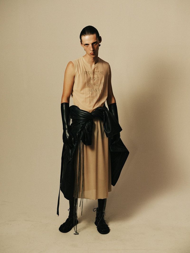 A slender person in a tan dress, black boots, and opera gloves stands against an off-white backdrop, a black jacket tied around their waist.