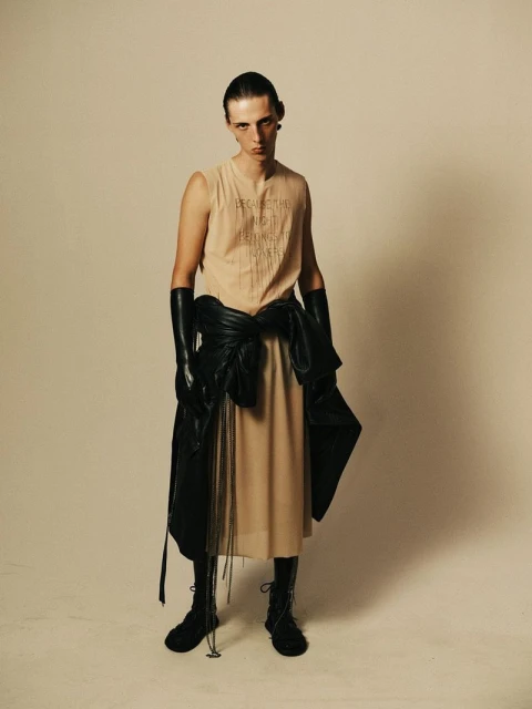 A slender person in a tan dress, black boots, and opera gloves stands against an off-white backdrop, a black jacket tied around their waist.
