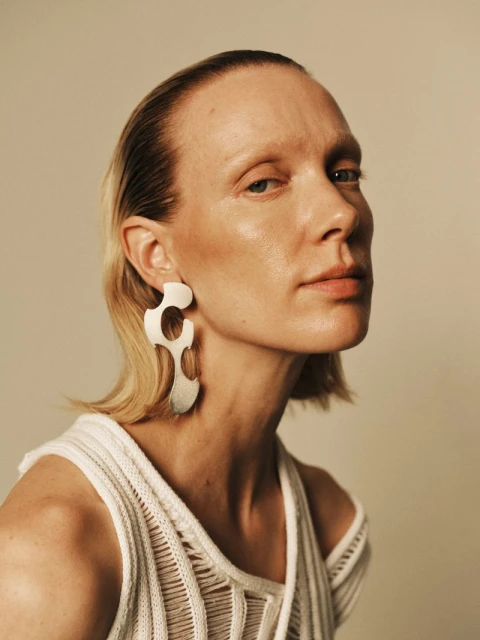Close-up portrait of an adult woman with short blonde hair wearing a white knit top and statement earrings, shot in a photo studio.