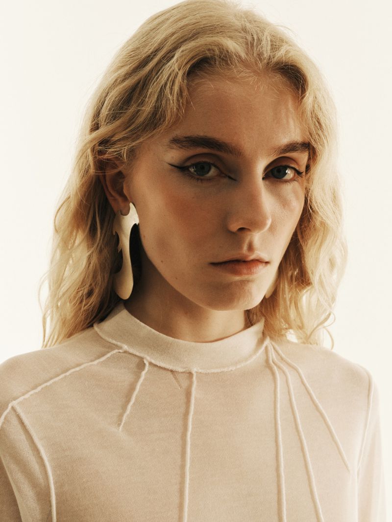 Close-up portrait of a young adult woman with long blonde hair, wearing a translucent top and large earrings, gazing at the camera.