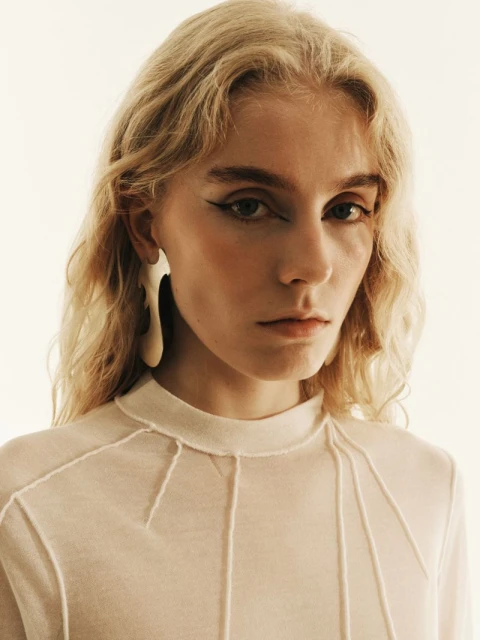 Close-up portrait of a young adult woman with long blonde hair, wearing a translucent top and large earrings, gazing at the camera.