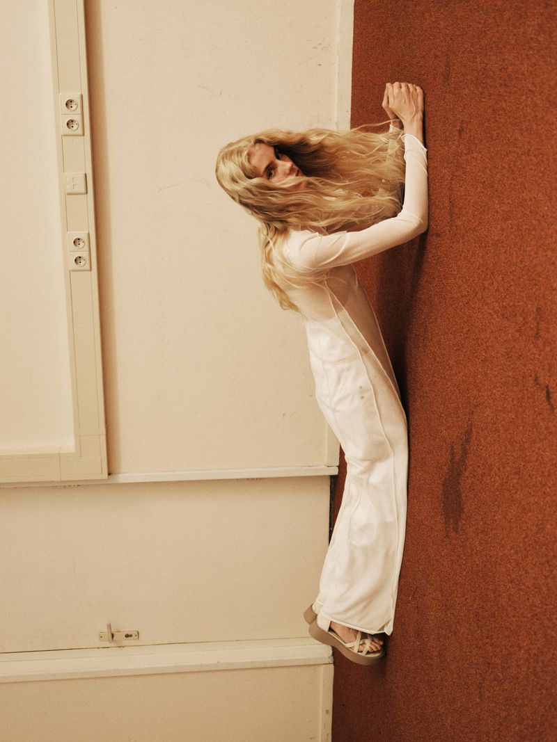 Blonde woman in a white outfit leans against a wall with brown and white surfaces. She wears wedge shoes and has a neutral facial expression.
