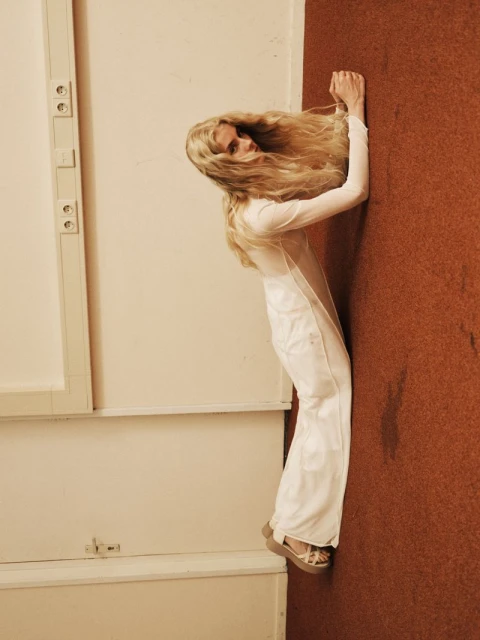 Blonde woman in a white outfit leans against a wall with brown and white surfaces. She wears wedge shoes and has a neutral facial expression.