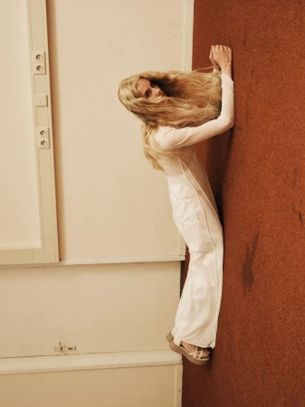 Blonde woman in a white outfit leans against a wall with brown and white surfaces. She wears wedge shoes and has a neutral facial expression.
