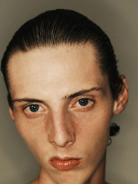 Close-up portrait of a young adult male with slicked-back dark hair, pale skin with freckles, and blue eyes against a neutral background.