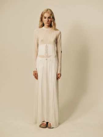 Model standing against a neutral studio backdrop wearing a sheer white long-sleeve top and matching long skirt.