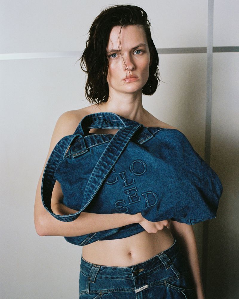 Fashion shot of a woman holding a denim bag in an indoor studio, featuring casual clothing and a neutral background, portrait orientation.
