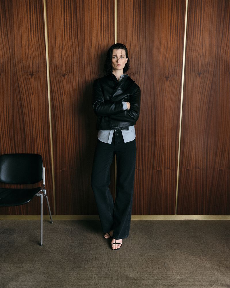 Full-length shot of a brunette woman with arms crossed, wearing a black leather jacket, dark jeans, and sandals against a paneled wall.