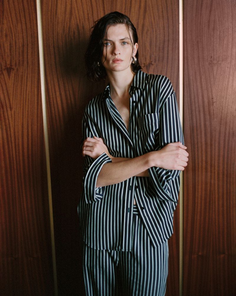 Adult woman poses indoors, wearing a vertically striped pajama-style black and white shirt and pants with a wood panel background.