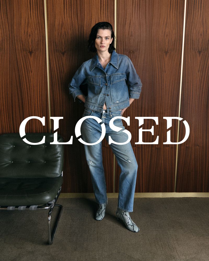 A woman poses indoors wearing a denim jacket and jeans with a wooden paneled wall behind her and a modern chair to the left.