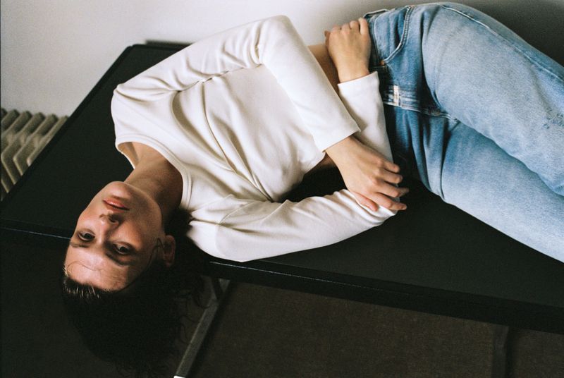 Adult woman with long dark hair reclines on black surface wearing white top and blue jeans in indoor environment with diffused lighting.