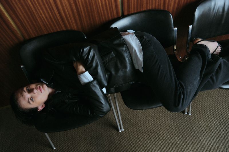 A woman in a black leather jacket reclines across three black chairs in an indoor setting, looking at the camera with a serious expression.