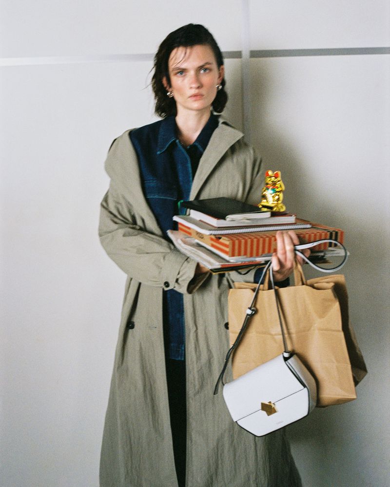 Woman in trench coat holds stack of notebooks and shoulder bag with a golden cat figurine on top against a white background.