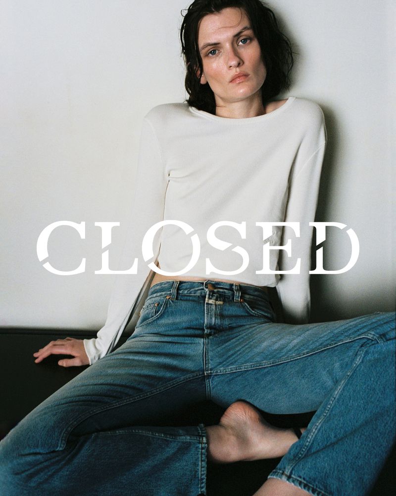 Indoor shot of a white adult female posing while wearing a long sleeve shirt and jeans, sitting on a couch with a white wall.