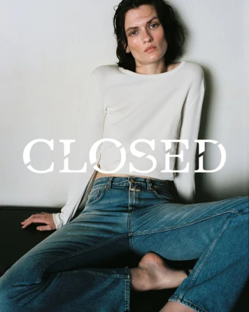 Indoor shot of a white adult female posing while wearing a long sleeve shirt and jeans, sitting on a couch with a white wall.