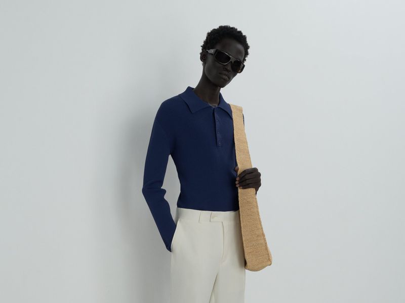 Dark-skinned adult model wearing a navy collared long-sleeved shirt with light beige pants and sunglasses in a studio setting.
