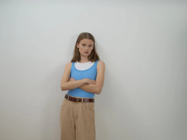 A woman with long brown hair stands in front of a white wall wearing a blue top, brown pants, and a brown belt while crossing her arms.