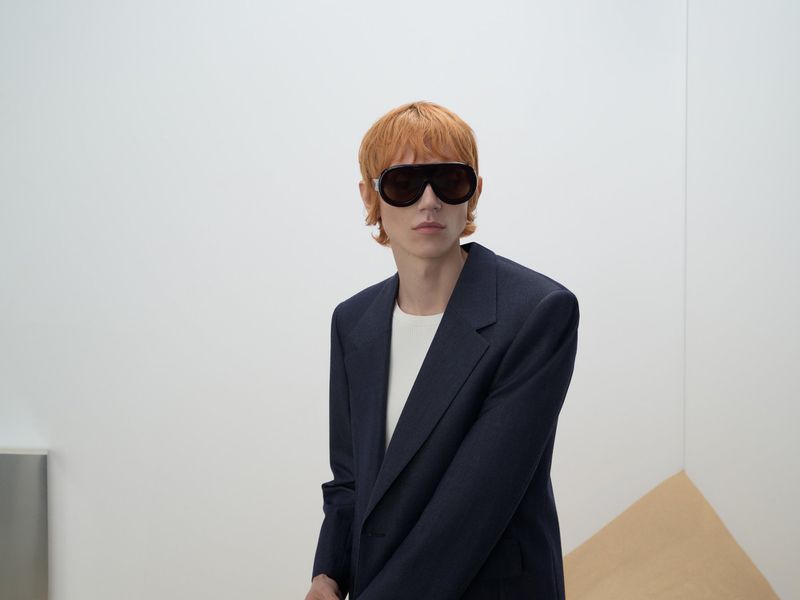 Model with orange hair and sunglasses wearing a dark blazer over a white shirt against a plain white background.