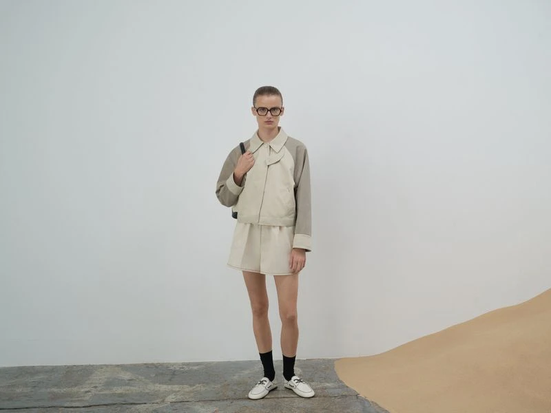 A woman in a neutral-toned jacket and shorts poses in front of a white wall with a tan design on the floor, wearing black socks and white shoes.